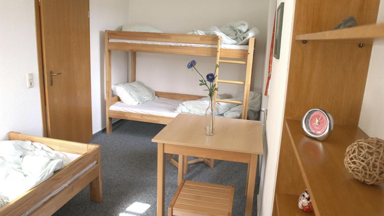 Een eenvoudige kamer met stapelbedden en een tafel in het midden. Aan de muur staat een kast met decoratieve voorwerpen.