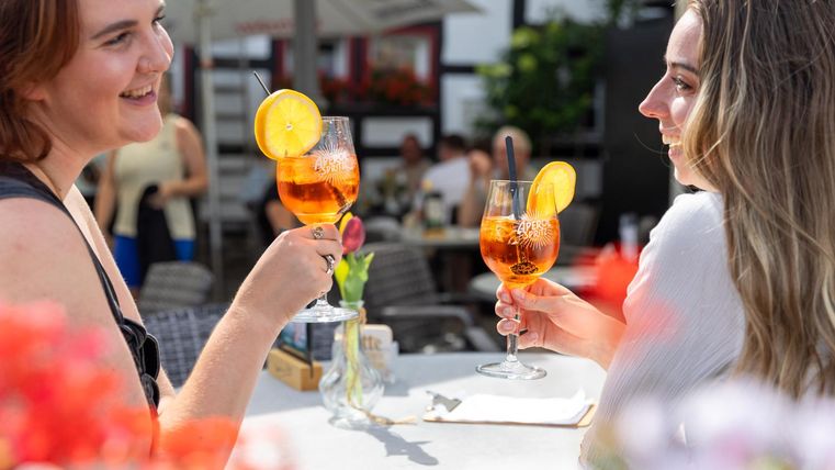 Two women toast with colorful drinks. In the background, there are people and blooming plants.