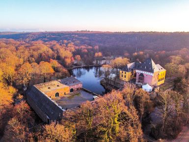 Ein malerisches Schloss am Rand eines klaren Sees, umgeben von buntem Herbstwald. Die Sonne beleuchtet die Szene in warmen Tönen.