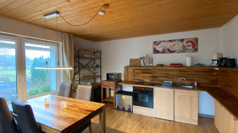 Eine moderne Küche mit Holzregalen und einem großen Esstisch mit fünf Stühlen. Viel Tageslicht fällt durch die Fenster auf die rustikale Einrichtung.
