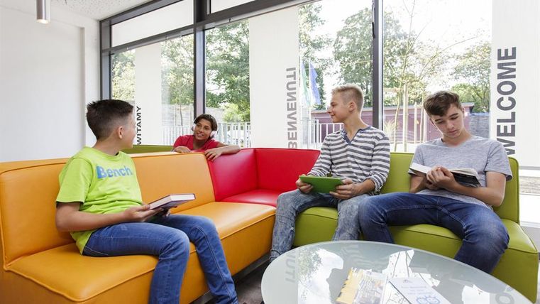 A group of four teenagers is sitting in a modern lounge. They are reading books and chatting while plenty of daylight streams through the windows.