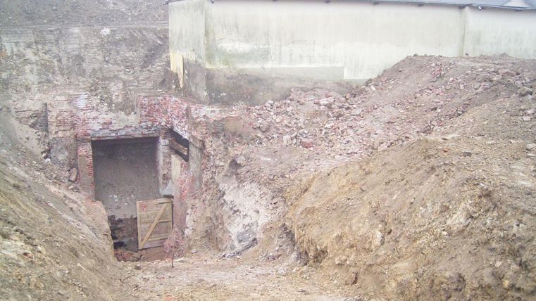 Eine Baustelle mit einem tiefen, leeren Graben und zerbrochenen Ziegelsteinen. Im Hintergrund ist eine graue Wand eines Gebäudes sichtbar.
