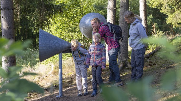 A family discovers an acoustic play device in the forest. The children listen attentively through the large, funnel-shaped hearing devices.