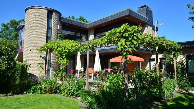 Ein modernes Haus mit großen Fenstern und einem grünen Garten. Im Vordergrund stehen Sonnenliegen und Sonnenschirme unter Weinreben.