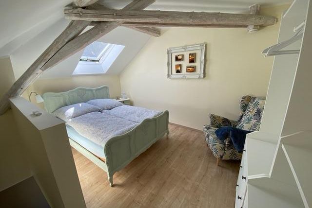 Een gezellige slaapkamer met een tweepersoonsbed en houten plafond. Er is een fauteuil en een tafeltje bij het raam.