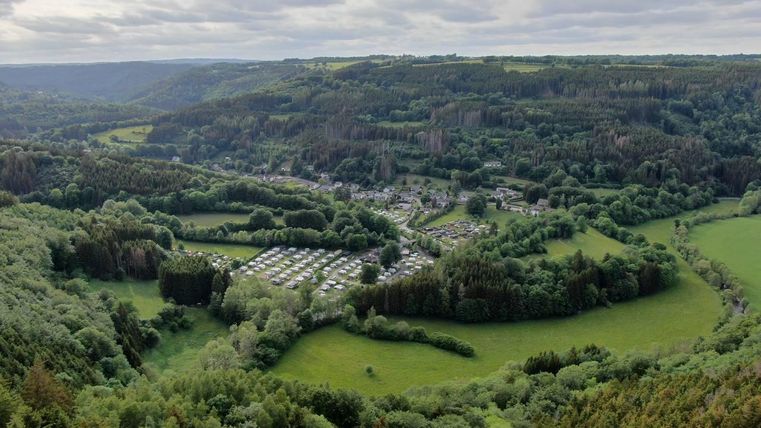 Een indrukwekkend landschap met groene heuvels en een klein dorp in de vallei. Op de voorgrond zijn campings en weilanden te zien, omringd door dichte bossen.