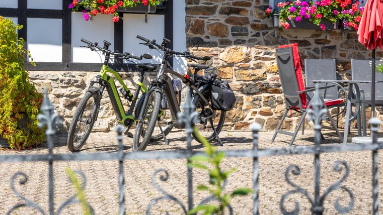 Twee fietsen staan naast een stenen muur bij een gezellige buitenruimte. Kleurrijke plantenbakken en parasols geven de scène een uitnodigende sfeer.