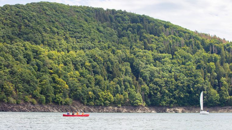 A tranquil lake surrounded by green hills. A red kayak and a sailboat are visible on the water.