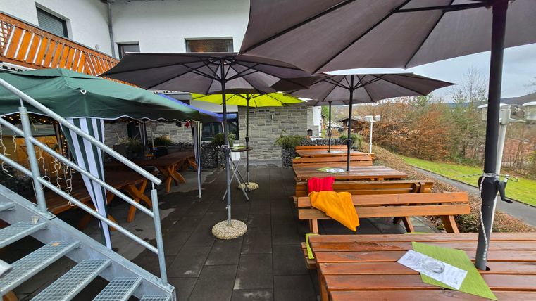A cozy outdoor terrace with wooden bench tables and large umbrellas. In the background, there are some green plants and a building visible.