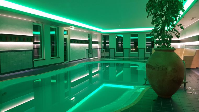 A modern indoor swimming pool with soft green lighting. In the background, large windows and a stylish plant can be seen.