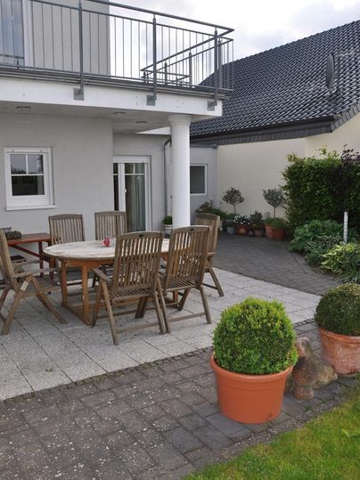 Ein gemütlicher Patio mit einem Holztisch und Stühlen. Pflanzkübel mit Pflanzen und ein modernes Haus im Hintergrund.