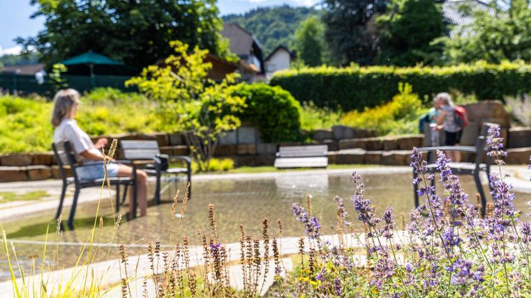 A quiet garden with a small pond and colorful flowers. In the background, two people are sitting on benches and enjoying nature.