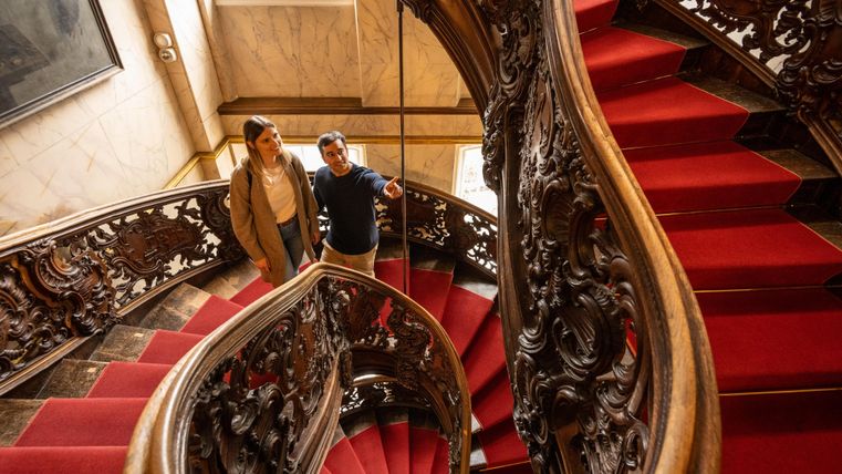 An elegant staircase with a red carpet and ornate wooden railing. Two people are standing and looking up.