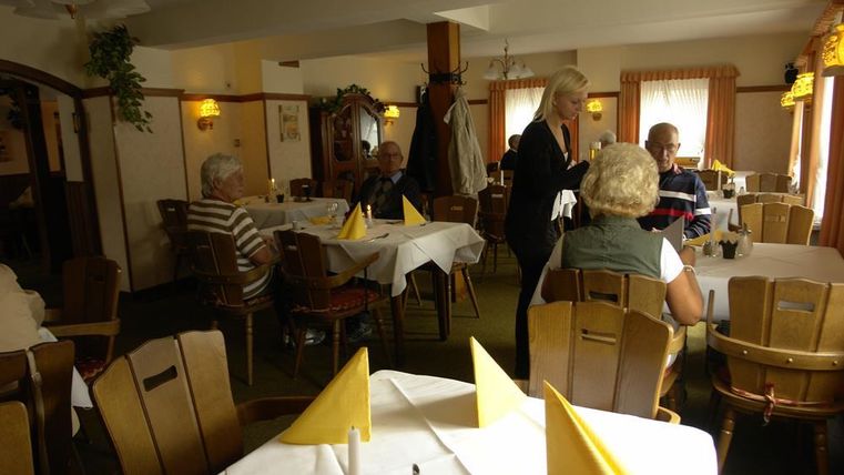 A cozy restaurant with tables dressed in white tablecloths and yellow napkins. Guests are sitting and chatting while a server takes their order.