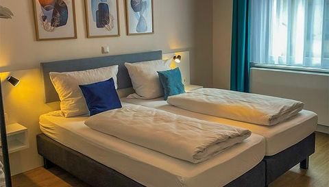 A cozy bedroom with two beds and bright pillows. Decorative pictures hang on the wall, and natural light floods through the window.