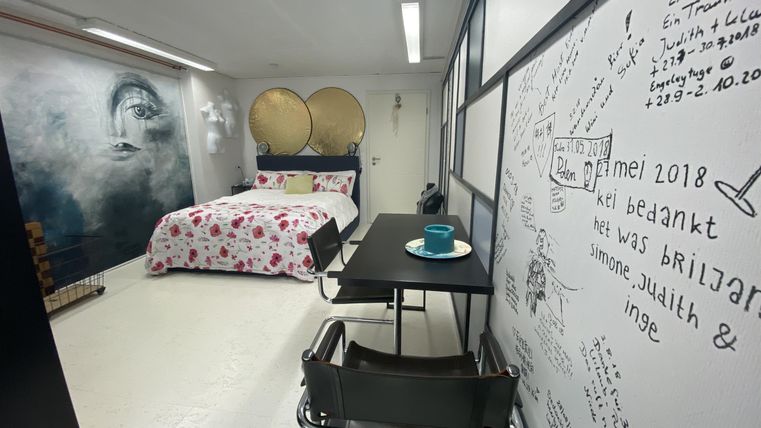 Een moderne kamer met een bed en een decoratieve wanddecoratie. Er is een tafel met een drankje en een creatieve muur met handgeschreven aantekeningen.