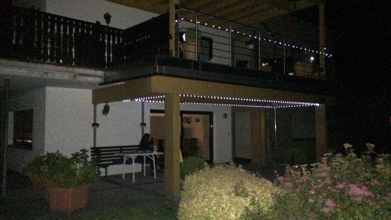 Ein Haus mit einem gemütlichen Balkon, der mit Lichtern dekoriert ist. Der Garten hat bunte Pflanzen und die Atmosphäre ist angenehm und einladend.