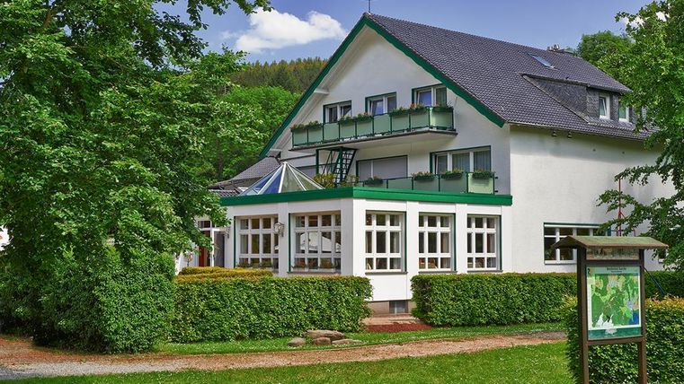 Ein charmantes Gebäude mit grünem Dach und Balkonen, umgeben von Bäumen und einer gepflegten Grünfläche. Der Himmel ist blau und klar, was eine einladende Atmosphäre schafft.