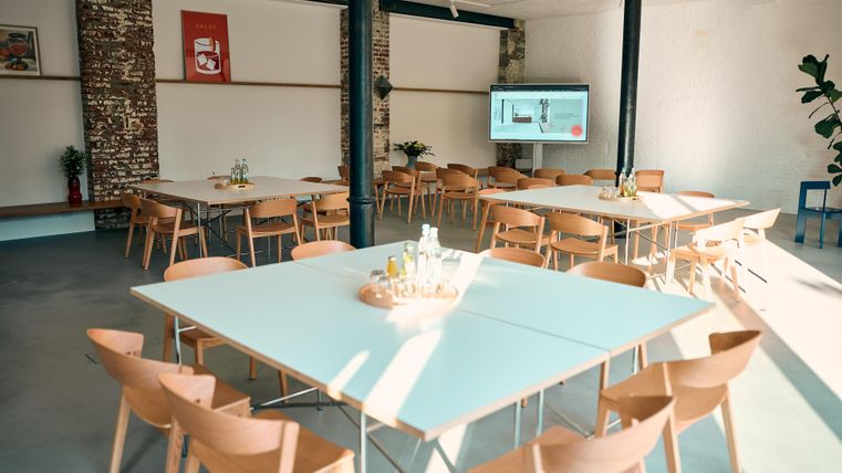 Ein moderner Konferenzraum mit mehreren Tischen und Stühlen. Ein Bildschirm ist an der Wand angebracht, und das Licht wirkt freundlich und einladend.