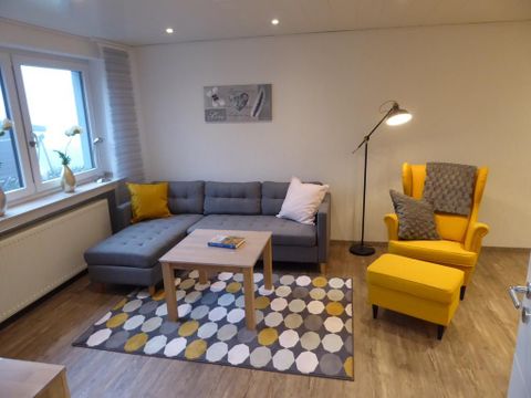 Een moderne woonkamer met een grijze bank en een gele fauteuil. De ruimte is helder en vriendelijk ingericht, met een tafel en een kleurrijk tapijt.