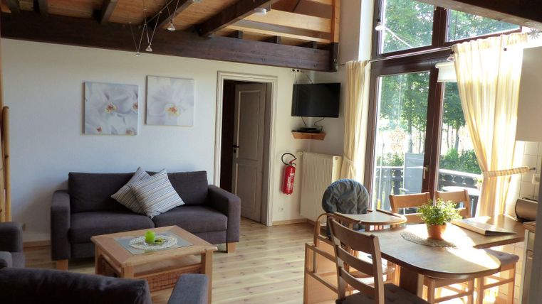 A bright living room with a comfortable couch and a dining table. Large windows provide a view of the green surroundings.