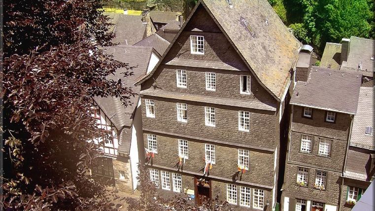Ein traditionelles, gut erhaltenes Fachwerkhaus in einer alten Stadt. Die Umgebung ist grün mit Bäumen und weiteren historischen Gebäuden.