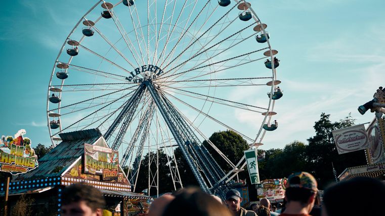 Ein Riesenrad steht auf einem Fest mit bunten Ständen und vielen Menschen. Der Himmel ist blau und wolkenlos.