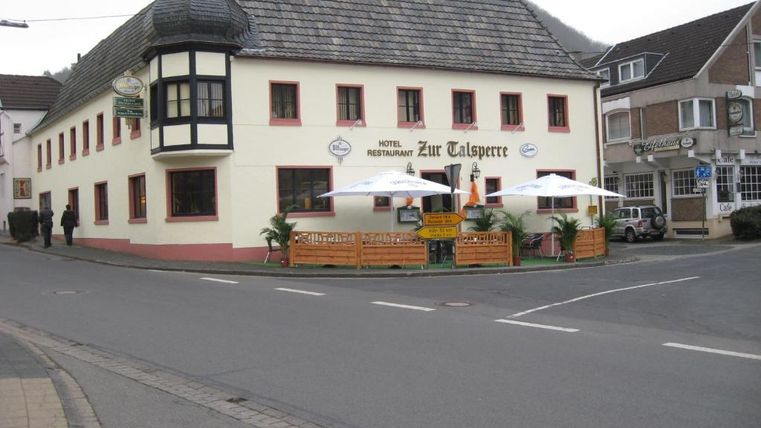 A charming restaurant and hotel named "Zur Talsperre" at a street corner. It has an inviting terrace with sun umbrellas and is surrounded by a quiet street.