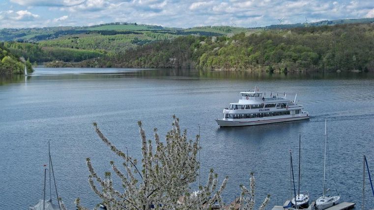 A calm lake with a boat sailing in the middle. In the background, green hills and a clear sky can be seen.