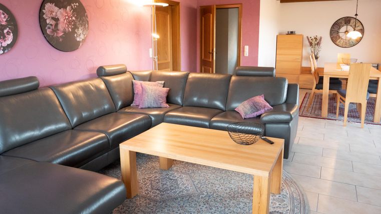 A modern living room with a large, comfortable couch and a wooden table. The walls are painted in a soft pink and decorative pictures adorn the space.