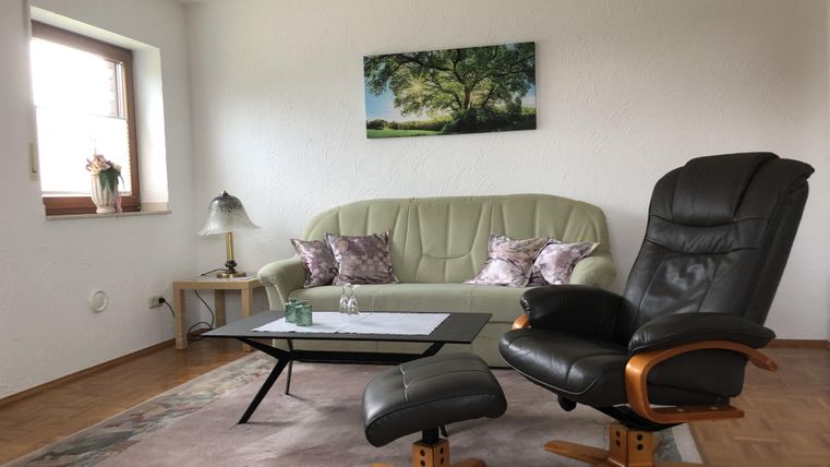 Ein gemütliches Wohnzimmer mit einem hellgrünen Sofa und einem schwarzen Relaxsessel. An der Wand hängt ein Bild von einem Baum, und der Raum hat Holzfußboden und eine kleine Lampe.