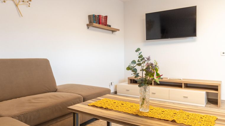 Een lichte woonkamer met een beige bank en een salontafel. Aan de muur hangt een televisie, en op de tafel staat een vaas met planten.