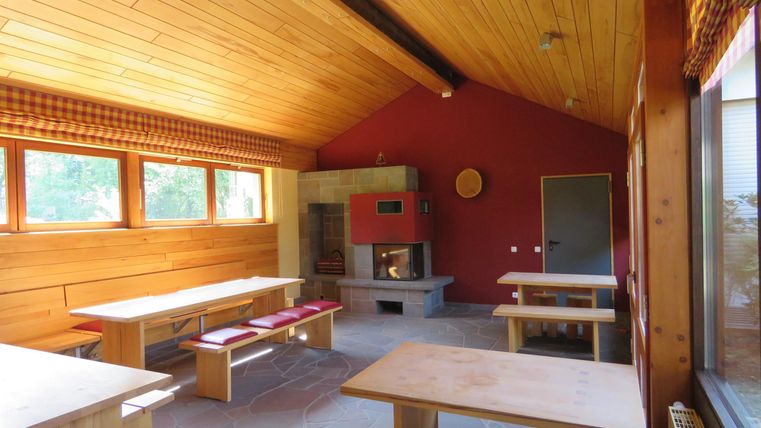 Ein heller Raum mit Holzwänden und Tischen aus Holz. Zu sehen ist auch ein Kamin und große Fenster, die viel Tageslicht hereinlassen.