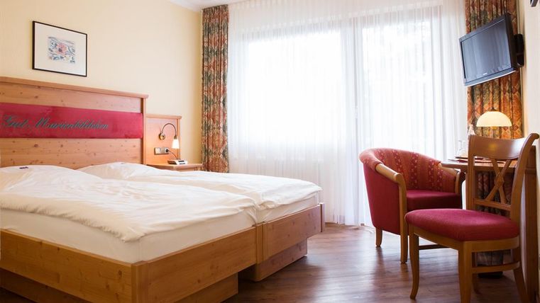 A cozy hotel room with two beds and wood interior. Large windows let in plenty of daylight, and there is a small workspace with a chair.