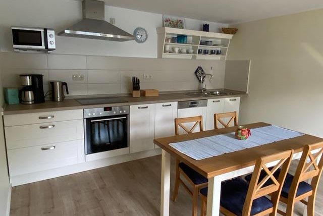 Een moderne keuken met lichte meubels en een grote eettafel. De uitrusting omvat een fornuis, een magnetron en verschillende keukenapparaten.