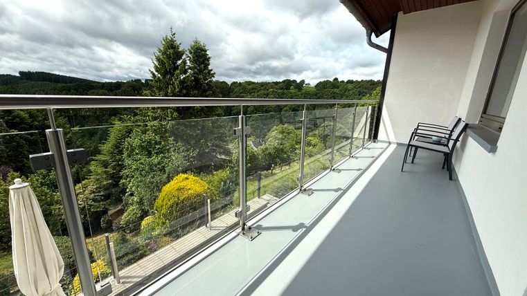 Een balkon met een glazen balustrade en uitzicht op het groene landschap. Er staan twee stoelen en een parasol aan de linkerkant.