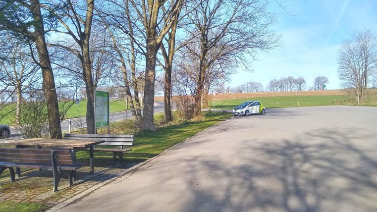 Een rustige parkeerplaats omringd door bomen en weilanden. De zon schijnt en de lucht is helder.