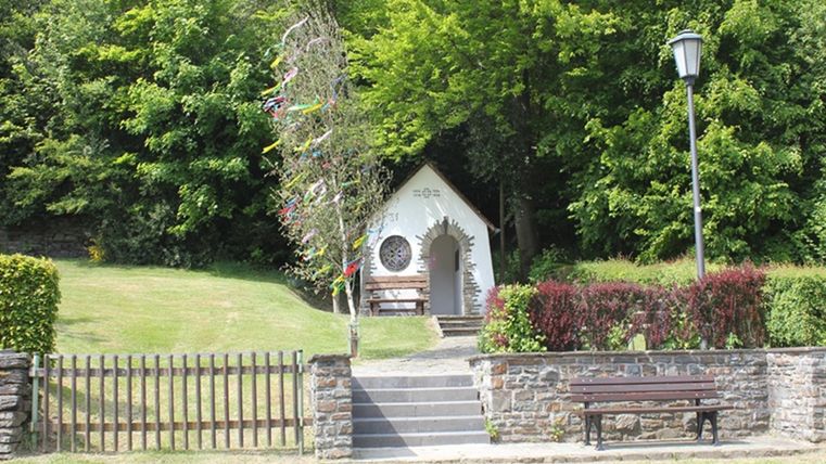 A small, charming little church stands surrounded by green trees. A bench in front of it invites you to linger.