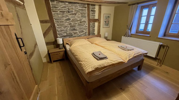 Een gezellige slaapkamer met een tweepersoonsbed en houten vloer. De muren zijn gedeeltelijk met steen en hout ingericht, en er zijn grote ramen die voor daglicht zorgen.