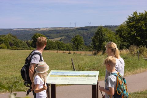 A family stands in front of an informational sign in a green landscape. In the background, rolling fields and trees can be seen under a clear sky.