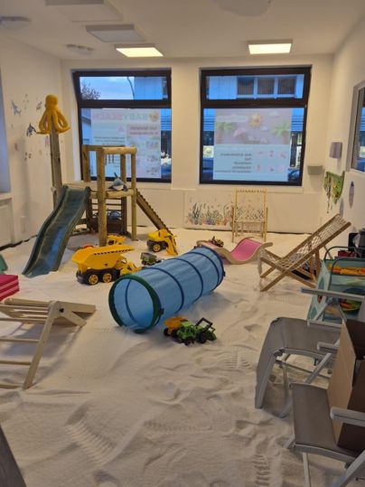 A playroom with a sand floor, toy cars, and a slide. In the center, there is a tunnel for playing and several seating options.