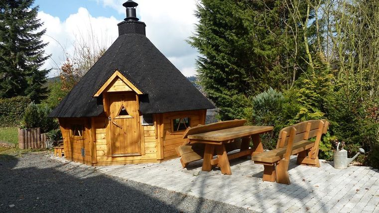 A cozy wooden cabin with a round roof and a wooden facade. Outside, there is a table set and some trees nearby.
