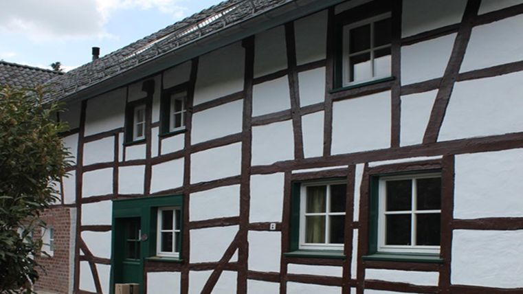 A traditional half-timbered house with white walls and brown beams. The windows are painted green and the roof is tiled.