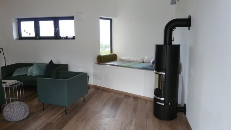 A modern living space with a cozy green couch and a wooden stove in the corner. Bright walls and large windows provide plenty of natural light.