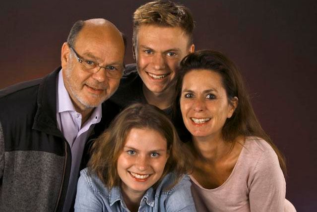 A happy family stands together and smiles at the camera. They consist of two adults and two teenagers.
