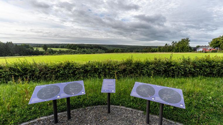 A viewing platform with information panels stands in front of a green landscape. The sky is cloudy and there is a distant view of the fields.