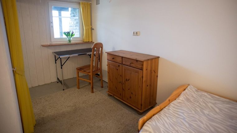 A simple room with a bed, a wooden desk, and a chest of drawers. Yellow curtains let daylight in.
