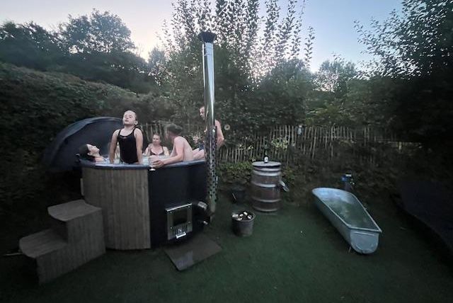 Een groep mensen ontspant in een buitenjacuzzi. Omgeven door groene bomen en een rustige tuinomgeving.
