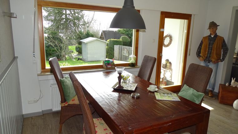 Een gezellige eetruimte met een grote houten tafel en sfeervolle verlichting. Door de ramen ziet men de tuin en een klein tuinhuisje.