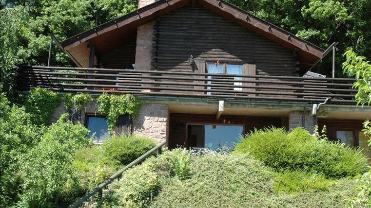 A beautiful wooden house with a balcony, surrounded by lush greenery. The garden and vegetation give the surroundings a tranquil atmosphere.
